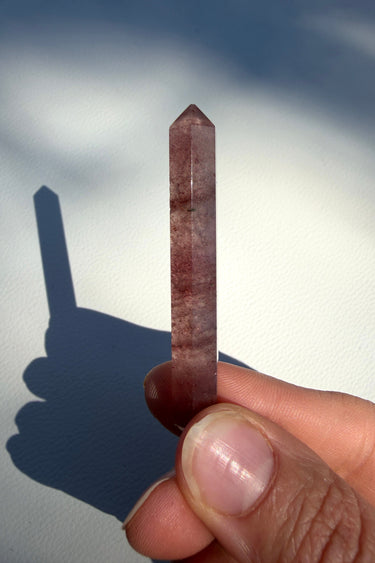 A polished berry quartz crystal point held between fingers, casting a sharp shadow on a white background.