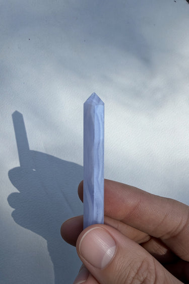 A hand holding a small pointed crystal or gemstone with a pale lavender-blue marbled pattern against a light gray background with a hand shadow.