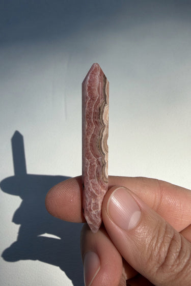 A hand holding a polished pink rhodochrosite crystal point against a light gray background with a prominent shadow.